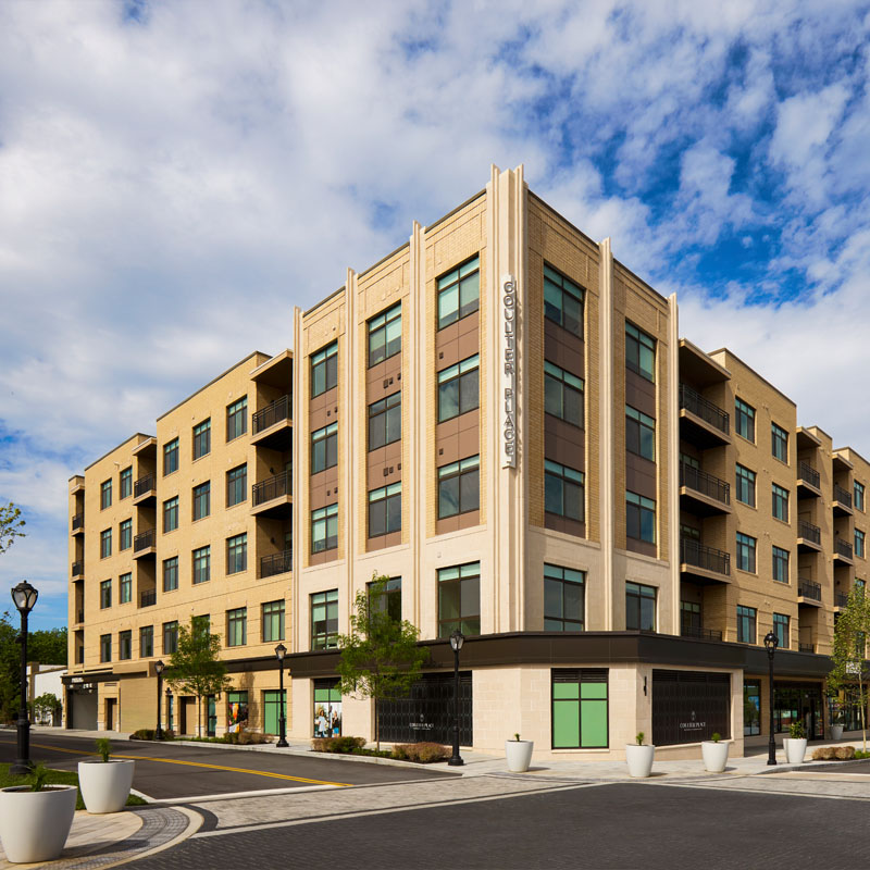 Street-level view of a Coutler Place residence with large windows, storefronts, and people walking and shopping along a landscaped sidewalk with trees and flowers.